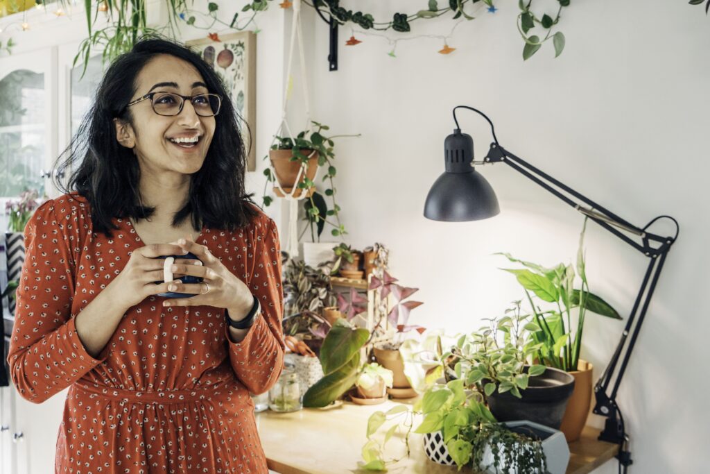 Lianne holding an enamel mug, in front of a table with houseplants and an angled lamp.